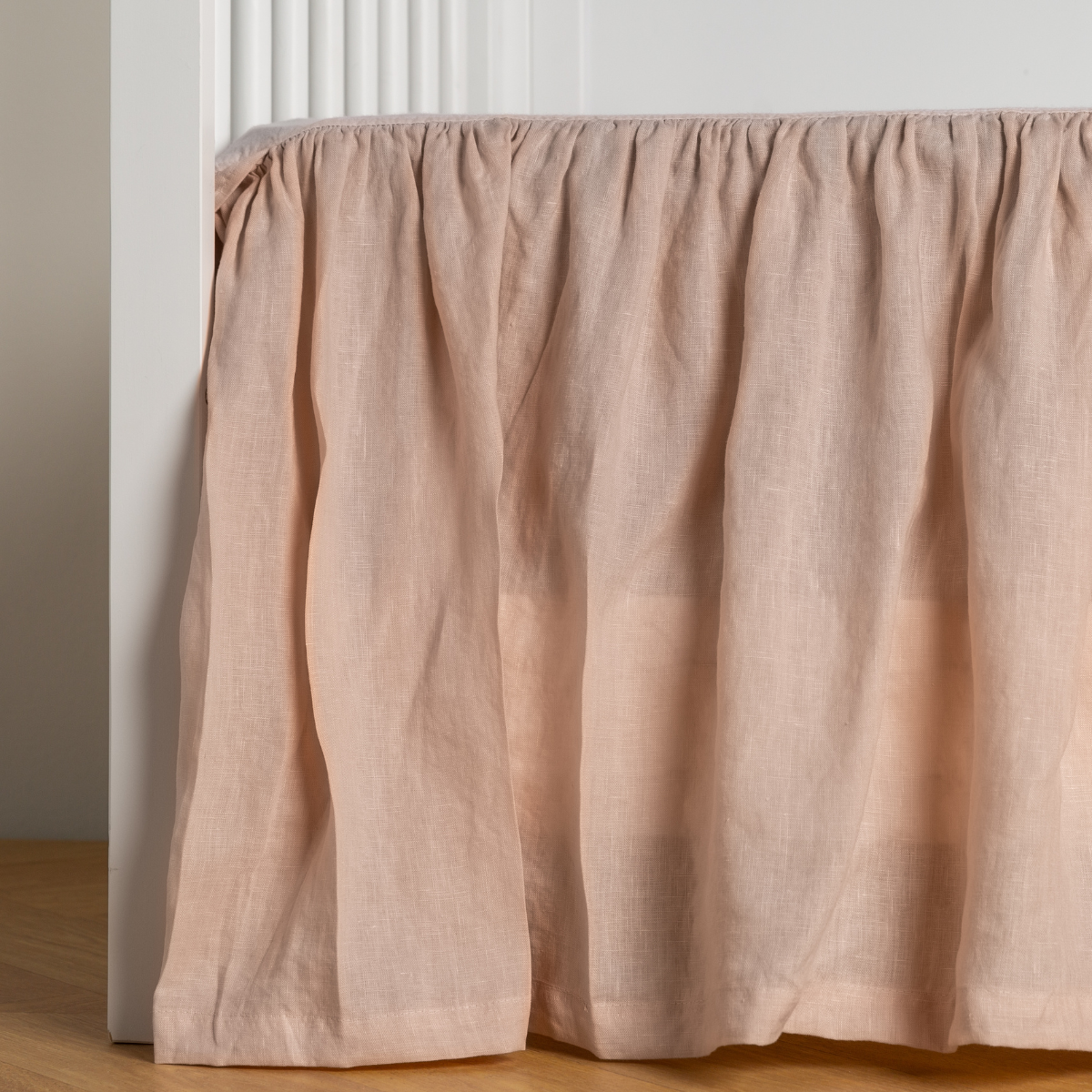 Gathered Pearl linen crib skirt draped to floor, side-angle view on light wood floor.