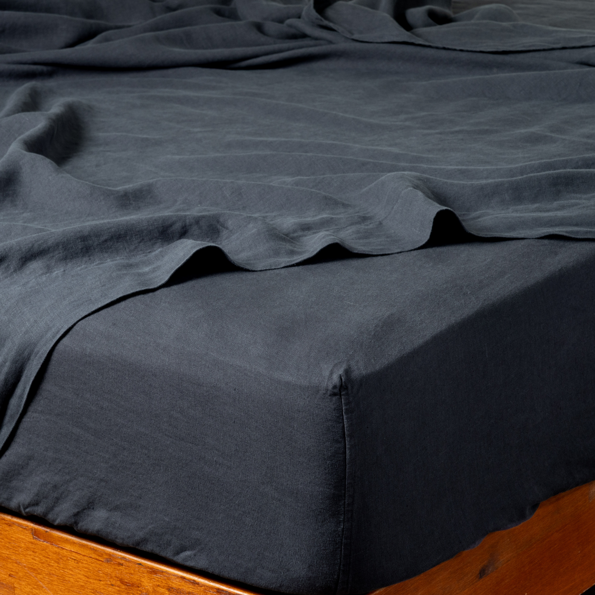 Corner detail of Bella Notte Linen fitted sheet in Slate, showing deep charcoal drape on wood bed frame.