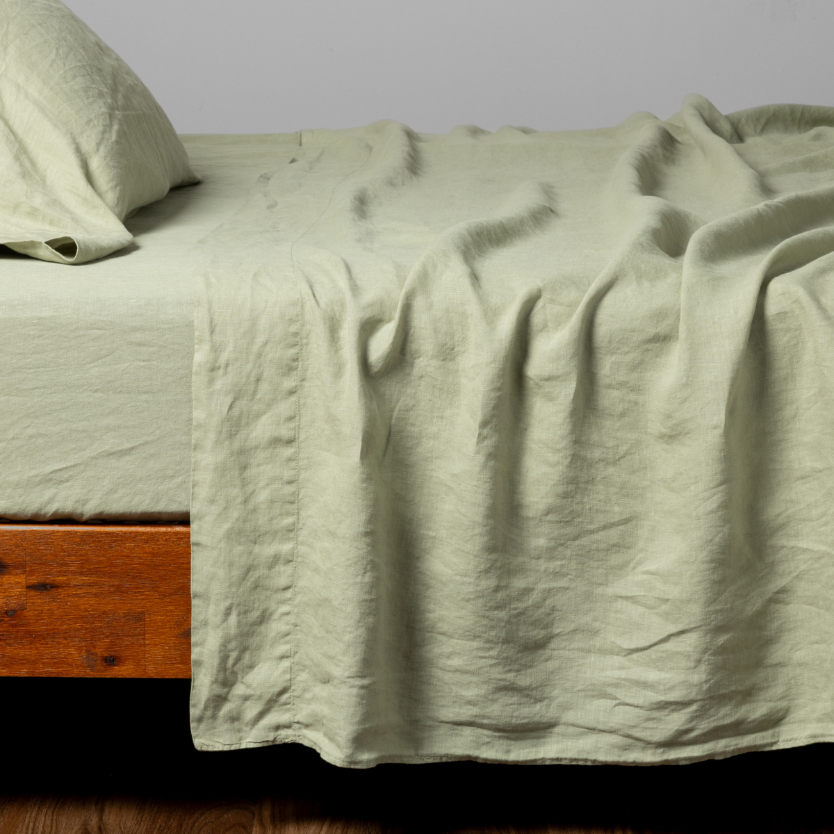 Desert Sage linen flat sheet draped over a wood bed frame, showing natural texture and relaxed drape.