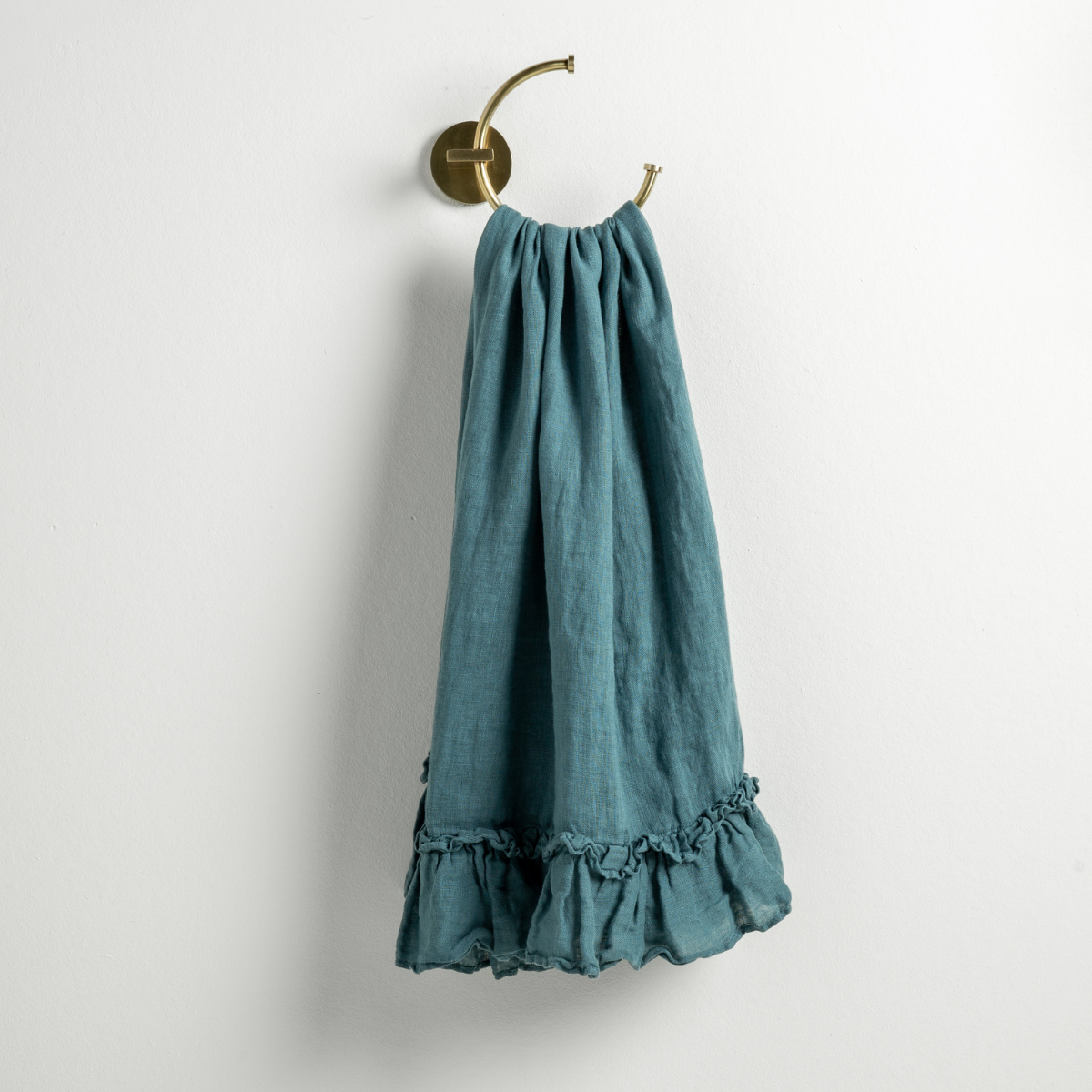 Cenote linen guest towel with ruffled hem, hung from a brass ring on a white wall.