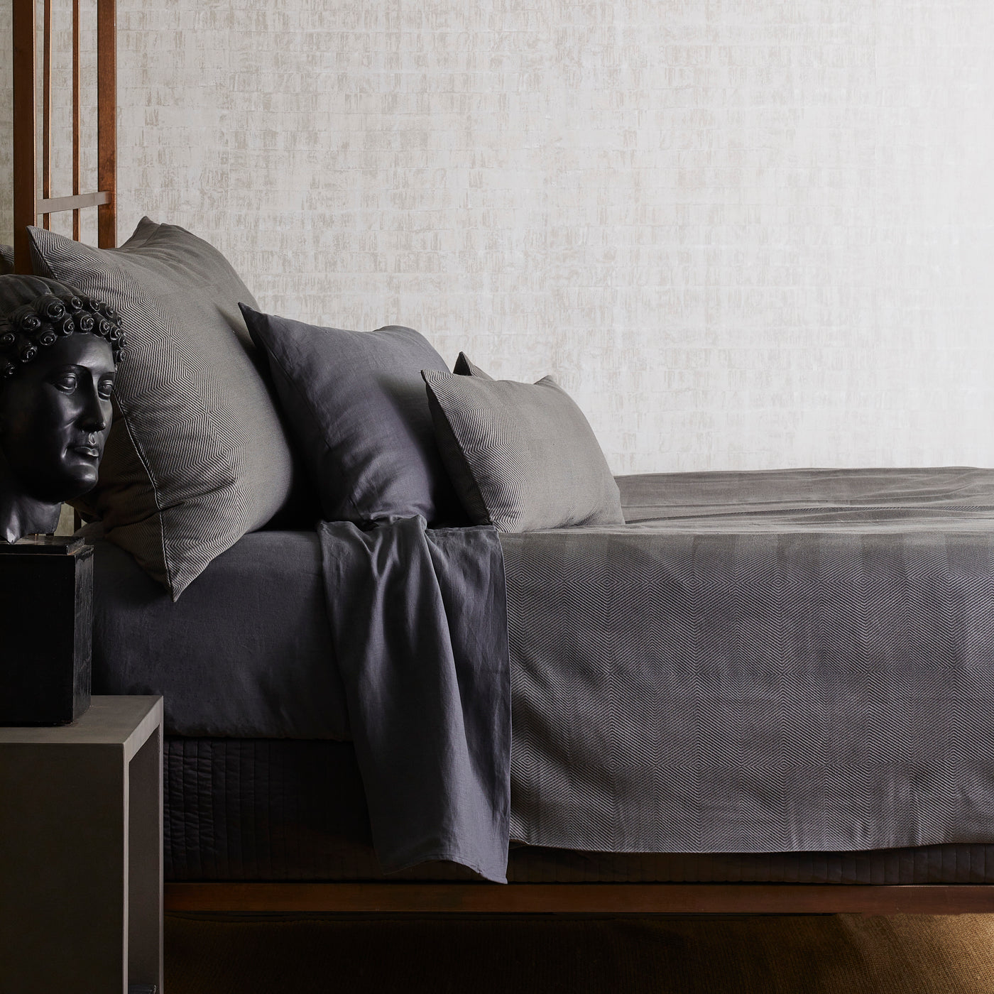 Charcoal linen sheet set draped over a wood bed frame, styled with a dark Buddha sculpture.