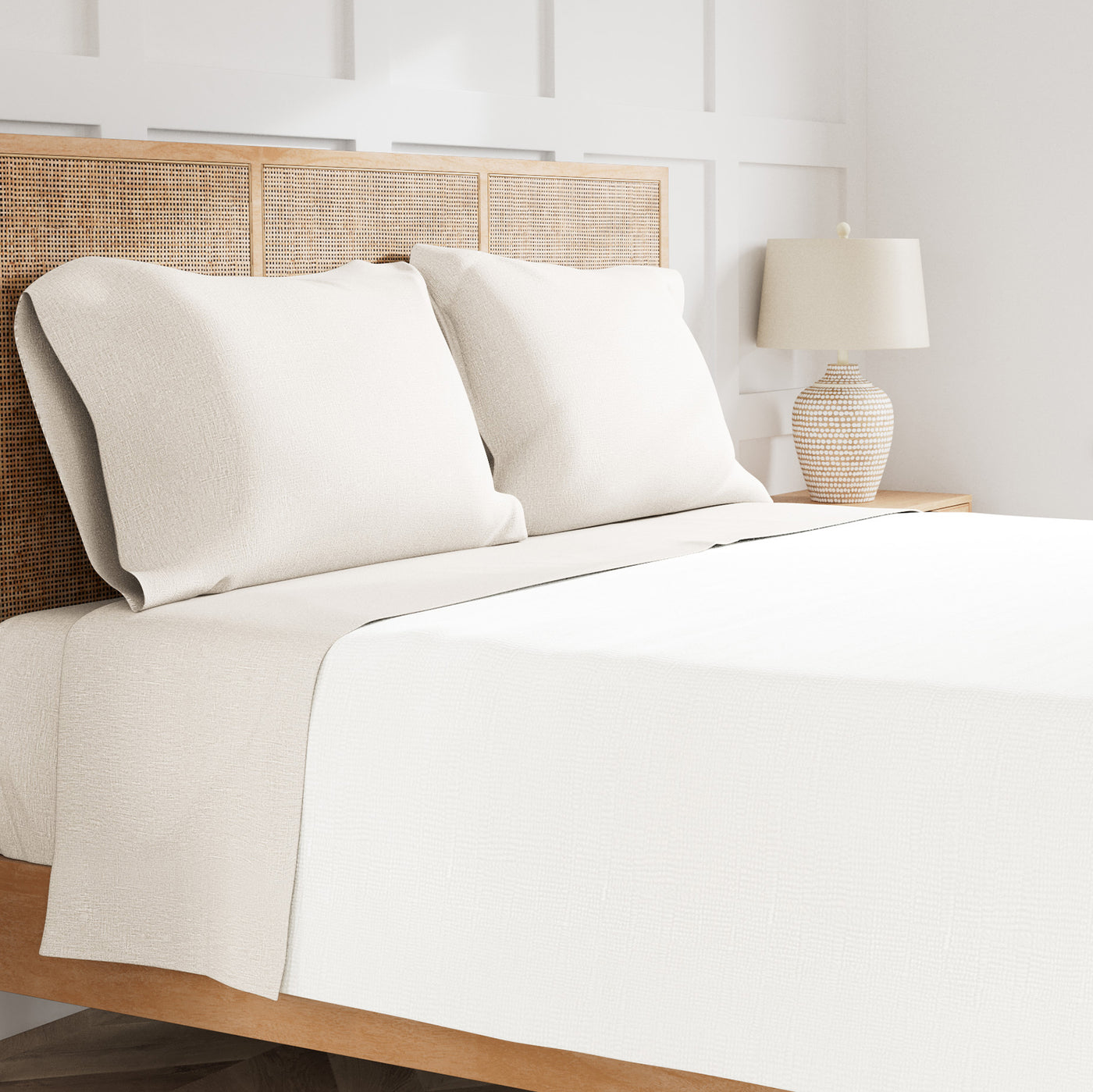 Ivory linen sheet set styled on bed with rattan headboard and neutral lamp accent.
