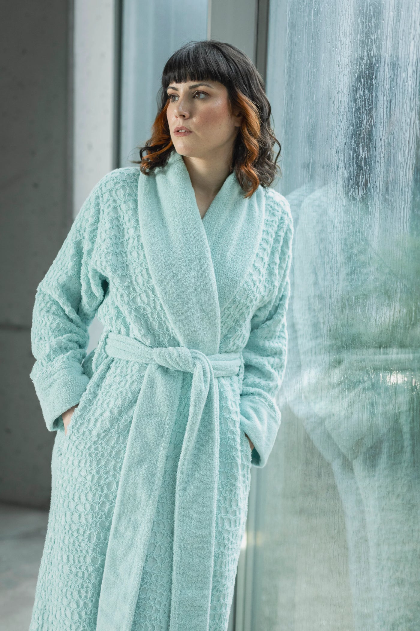 Model wearing Abyss Habidecor Lodge robe in aqua, full-length view beside glass wall.