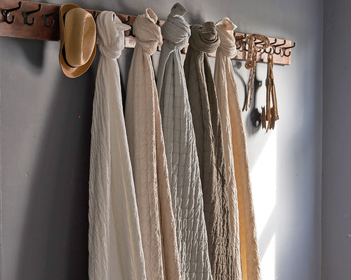 Multiple Traditions Linens Louisa coverlets in neutral tones draped from a wall-mounted coat rack.