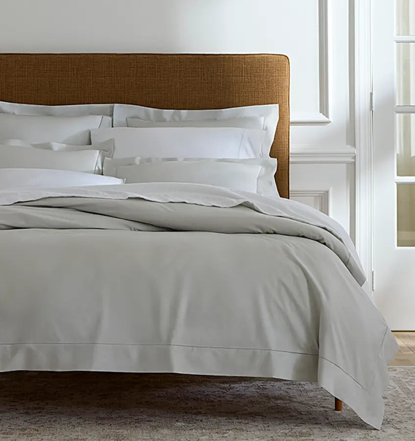 Sferra Luxor duvet cover in silver-grey draped over a made bed with woven headboard.