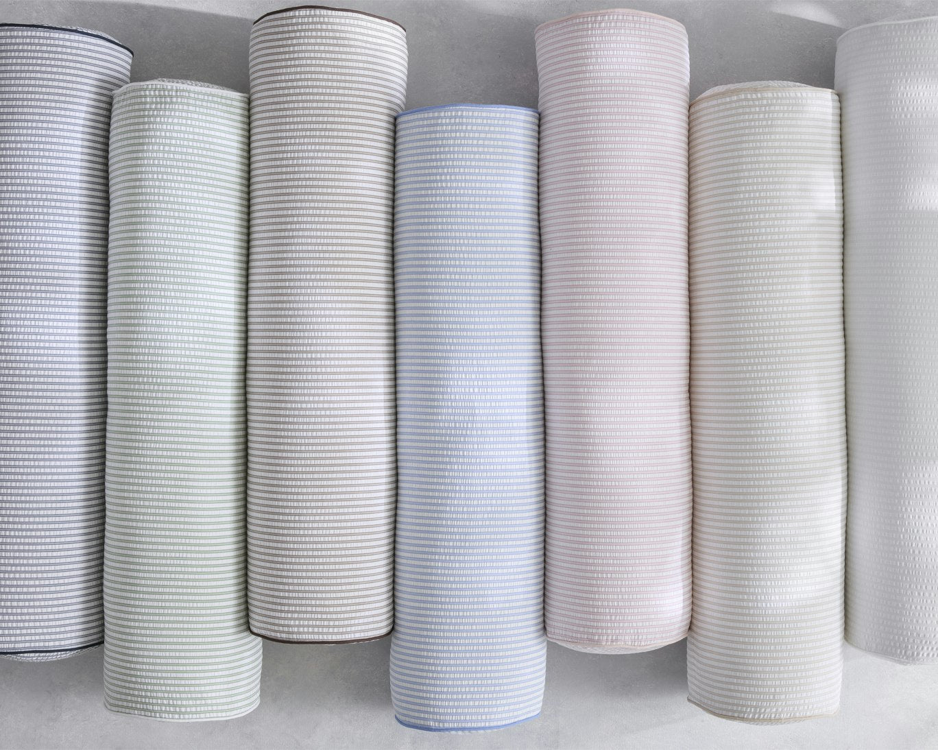 Six rolls of textured fabric in various light colors, including gray, white, and pale blue, arranged vertically against a neutral light gray background.