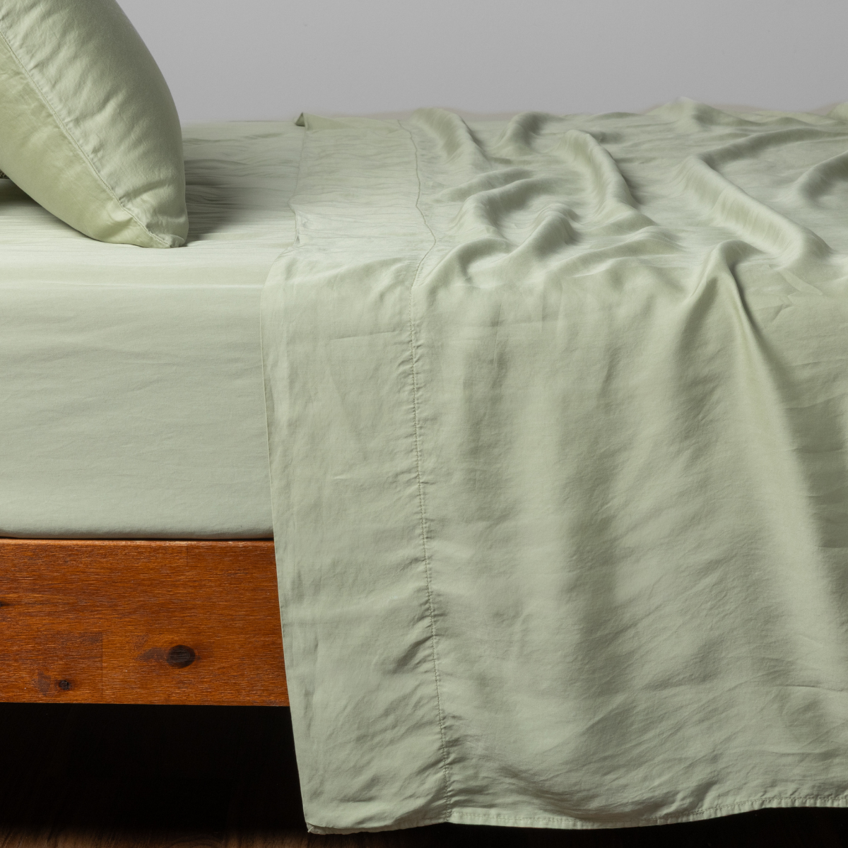 Madera Luxe flat sheet in Desert Sage, casually draped over a wood-framed bed.