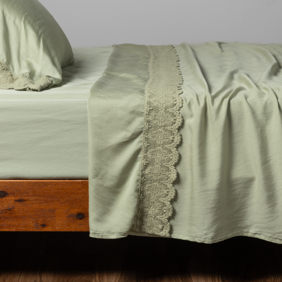 Desert Sage flat sheet with wide scalloped lace trim, draped over a wood-frame bed.