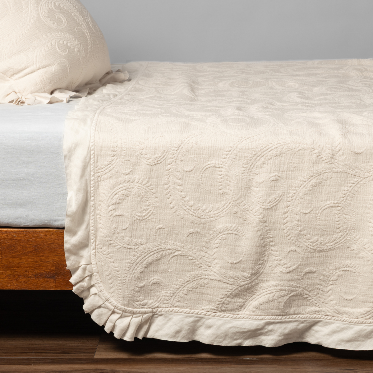 Natural quilted bedspread with swirling matelassé pattern and ruffled hem, draped over bed corner.