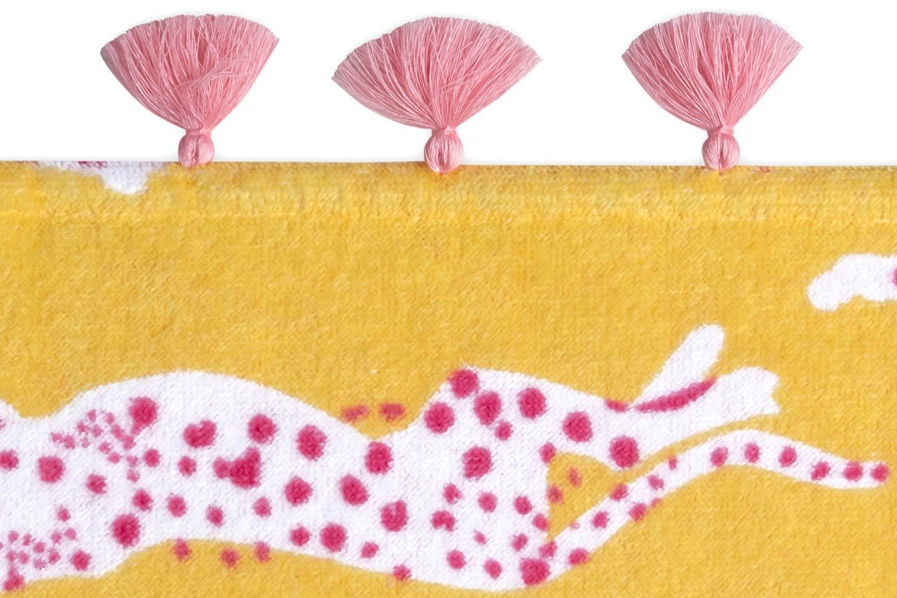 The Matouk Schumacher Leaping Leopard Beach Towel is crafted from yellow cotton velour with a white and pink spotted animal pattern and three pink tassels along the top—playful and stylish for your beach days.