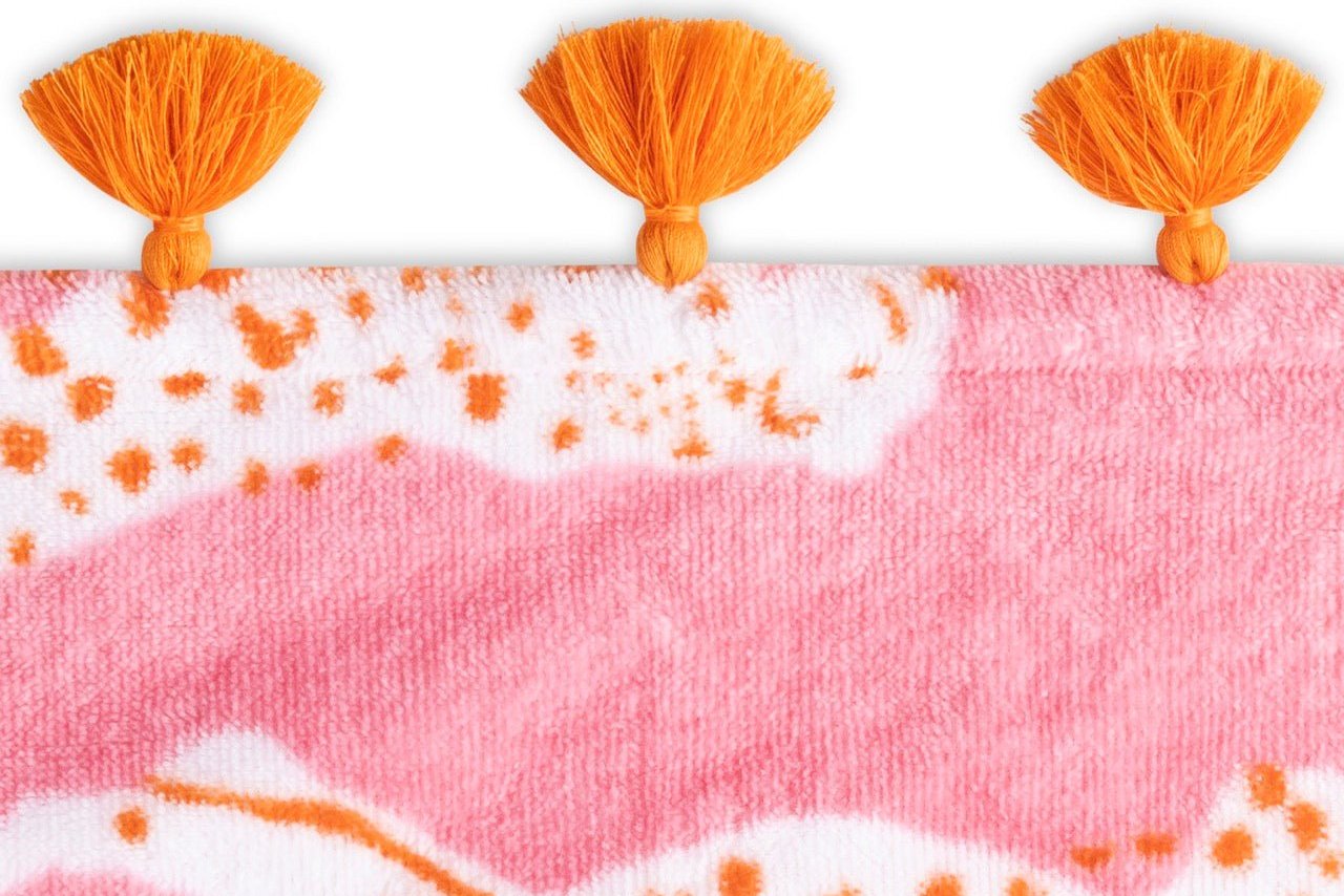 Close-up of the Schumacher Leaping Leopard beach towel by Matouk, featuring pink and white patterned cotton velour with three orange tassels along the top edge.