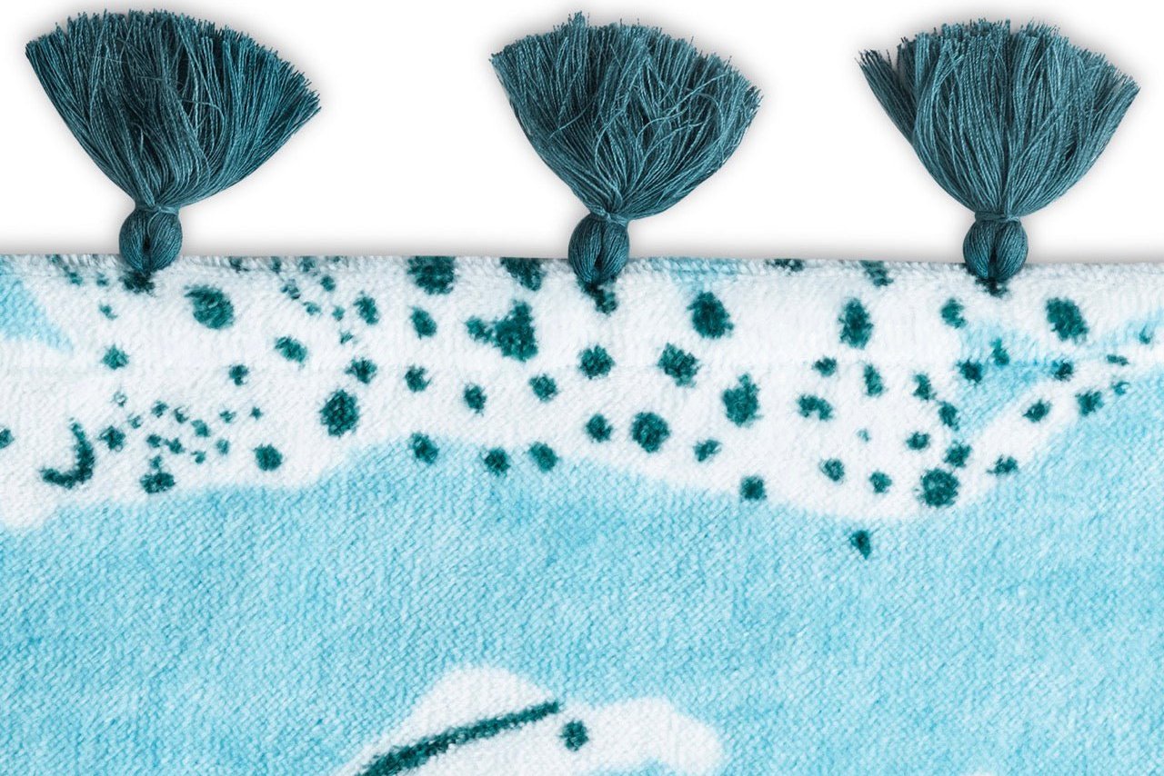 A close-up of the Schumacher Leaping Leopard beach towel by Matouk, featuring blue and white patterned cotton velour and three teal tassels along the top edge—perfect for poolside lounging.
