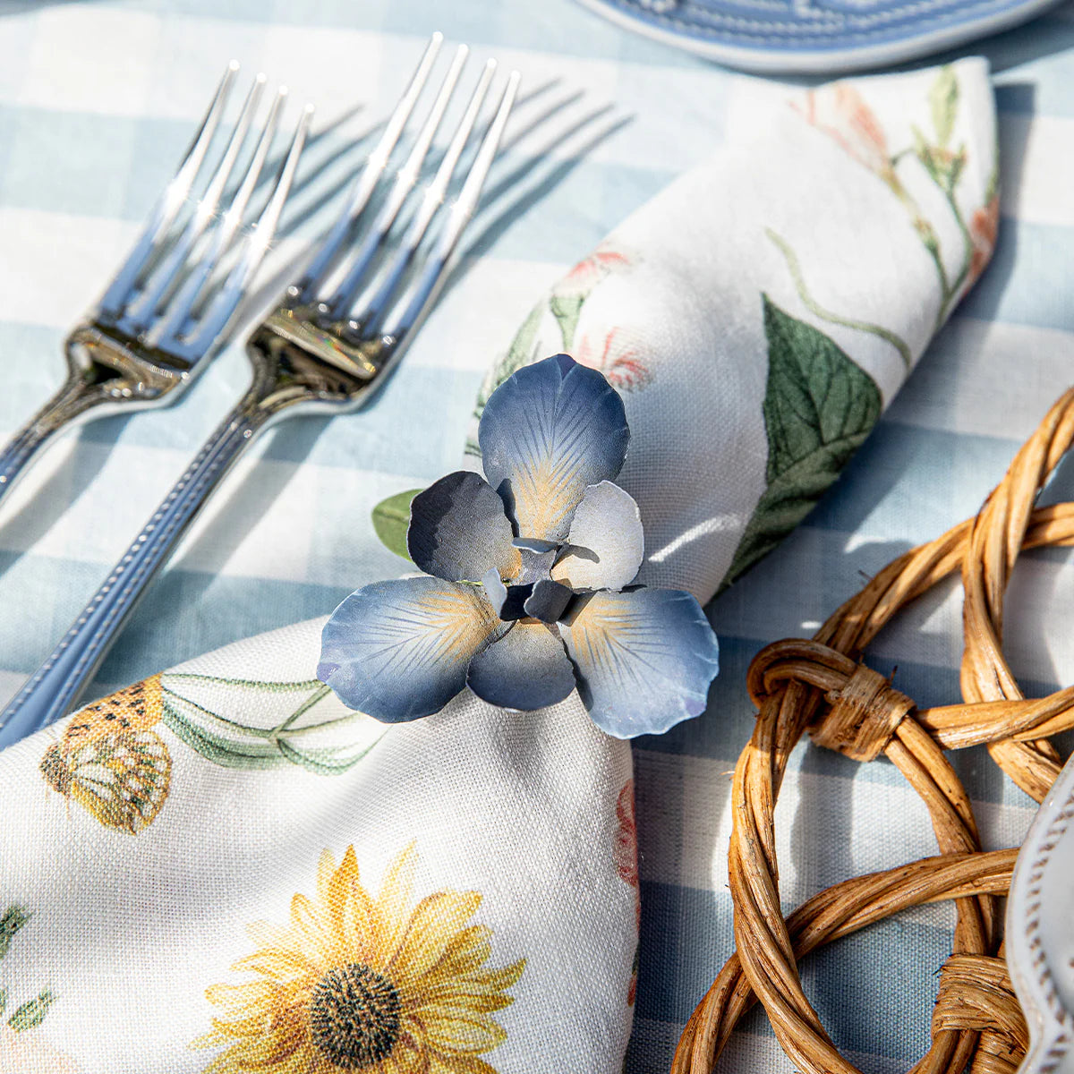 Blue iris flower napkin ring holding a floral linen napkin on a gingham tablecloth.