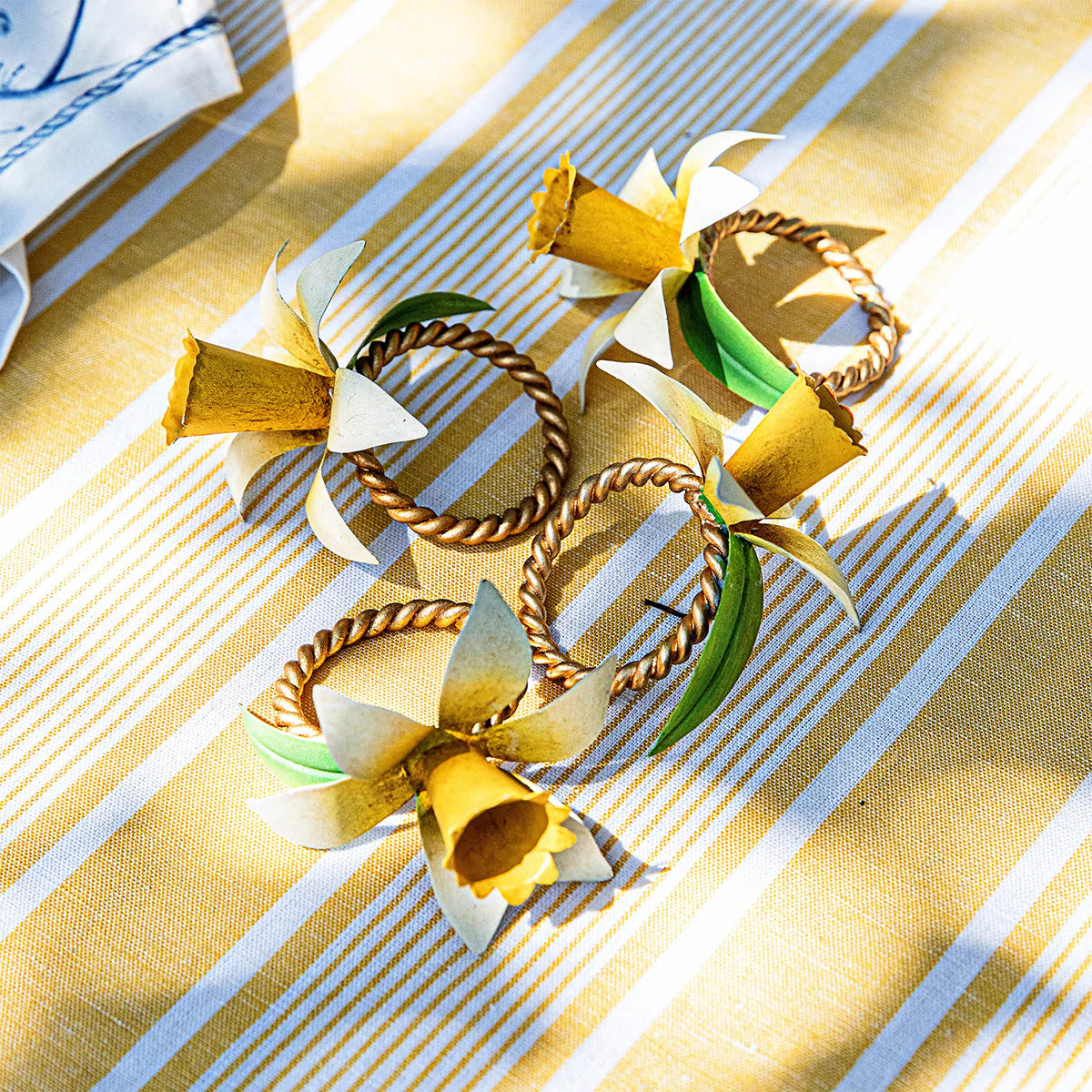 Four Juliska Meadow Walk napkin rings with daffodil blooms on a gold-striped linen tablecloth.