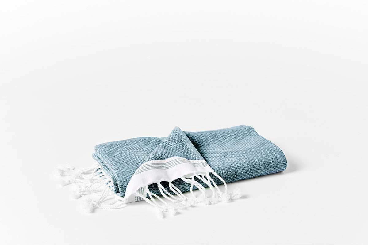Folded teal waffle-weave towel with white knotted fringe, styled flat on white surface.