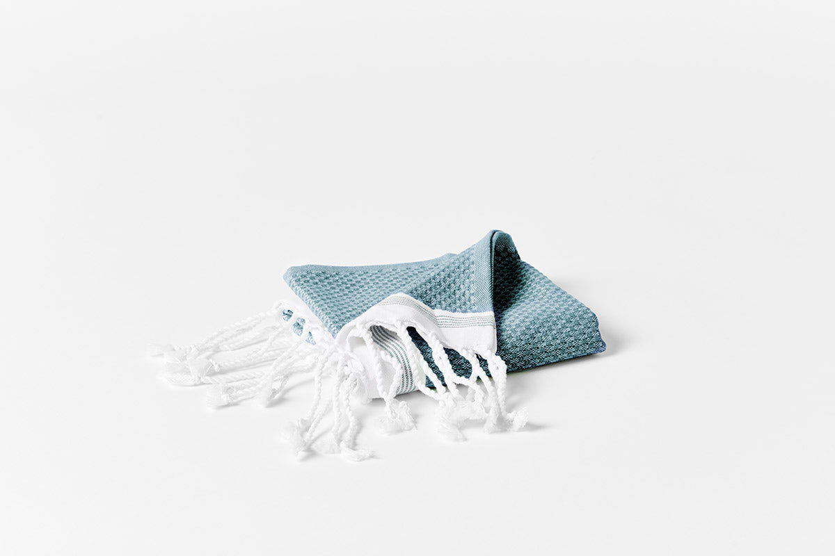 Folded teal waffle-weave bath towel with white fringe, shown on white surface.