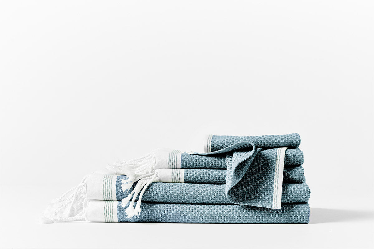 Coyuchi Mediterranean towel set stacked, slate blue waffle weave with white fringe and stripe trim.