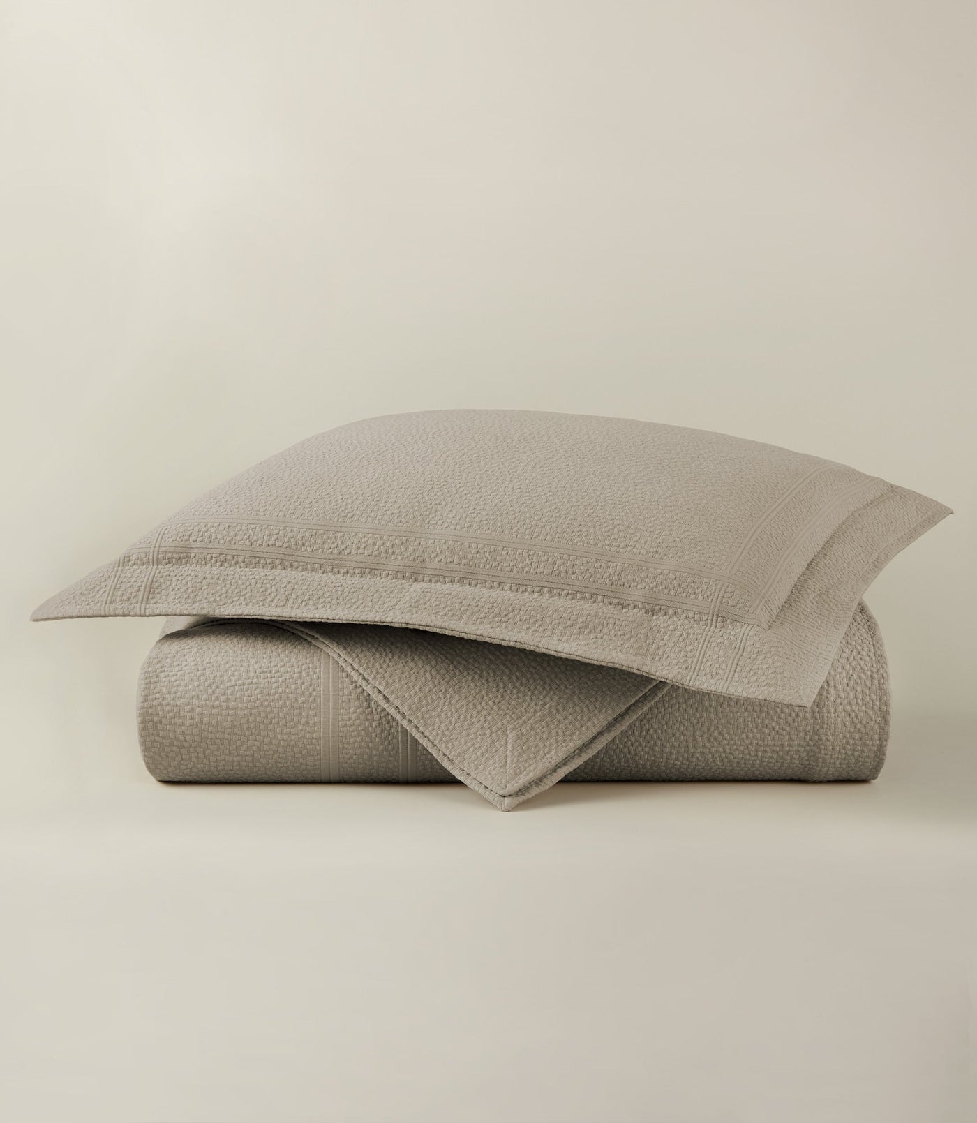 Folded taupe matelassé coverlet and matching sham stacked against a neutral background.