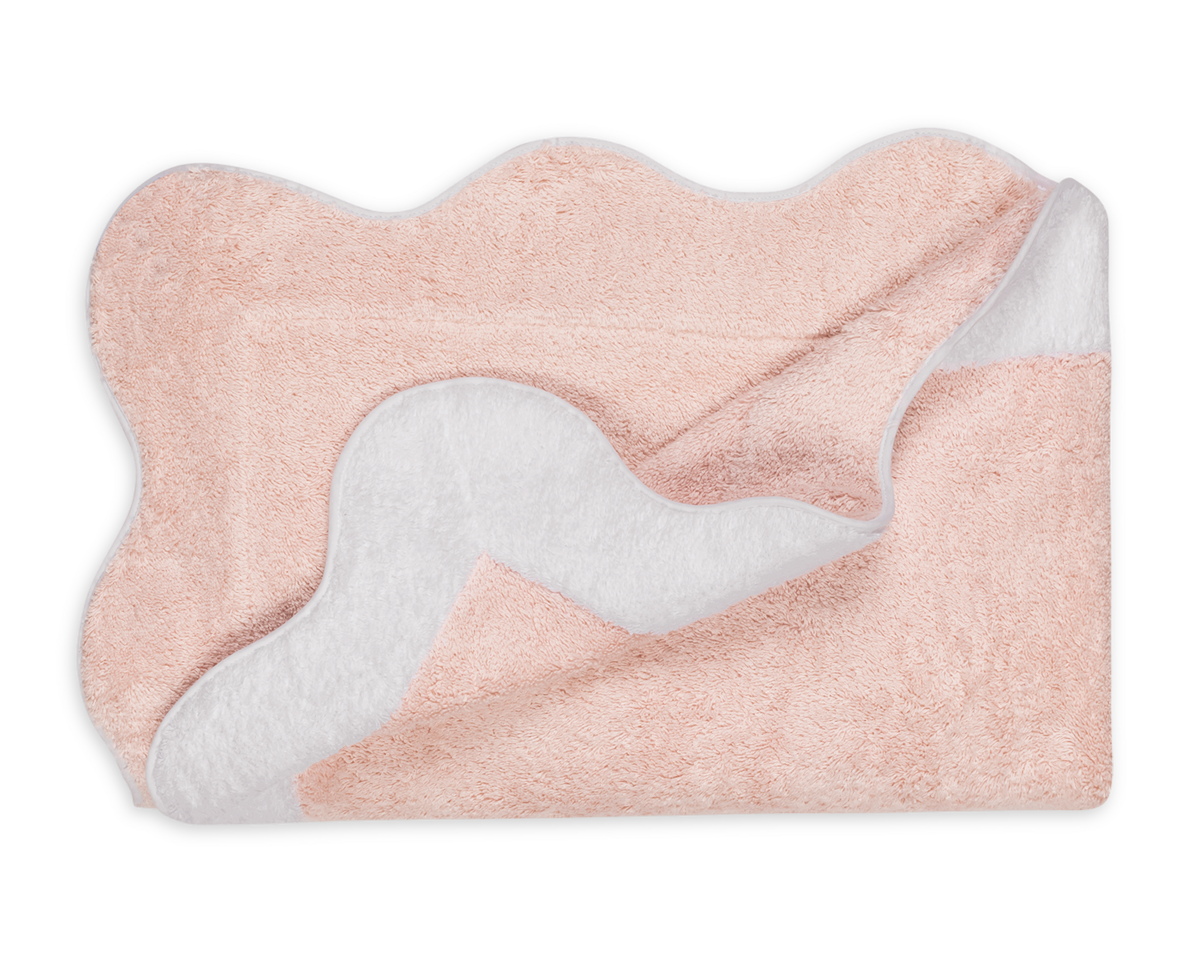 The Neptune | Pool Towel by Matouk is a soft, light pink rectangular towel with wavy edges and a white underside, shown partially folded to display both sides.