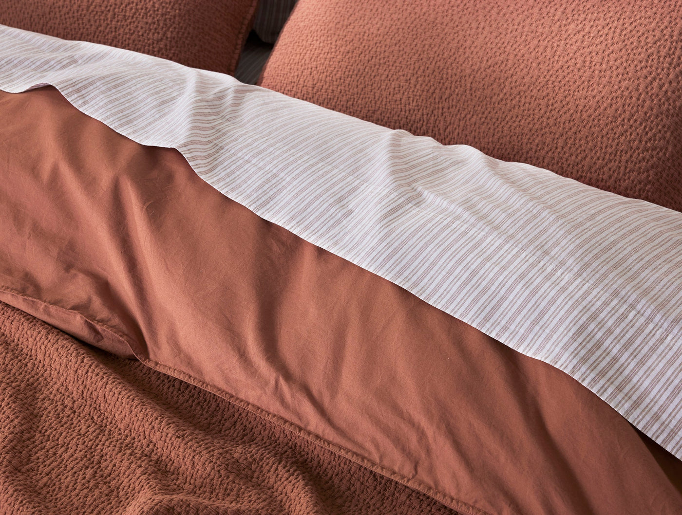 Close-up of white striped Coyuchi Crinkled Percale pillowcase folded over terracotta bedding.