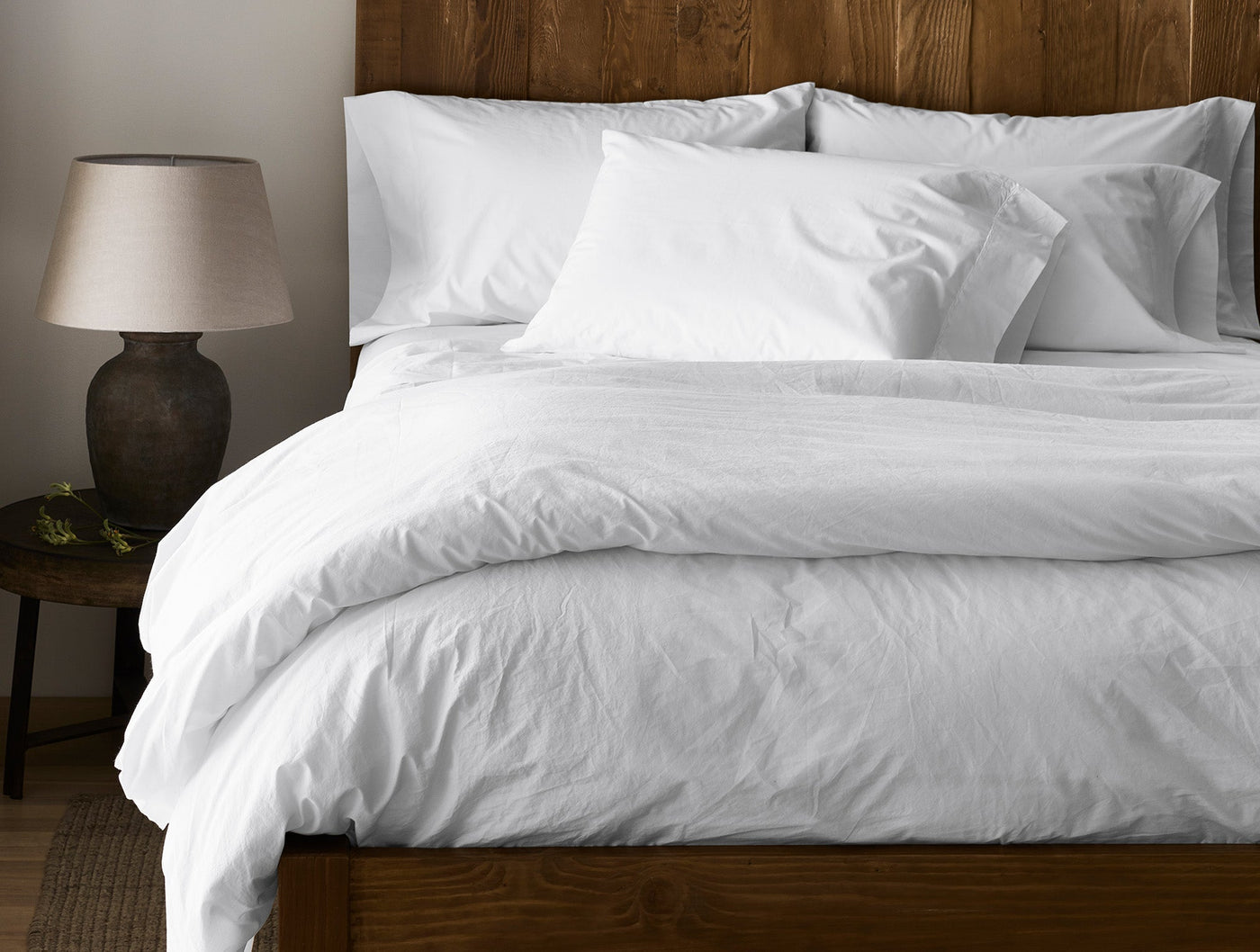 Softly rumpled white percale bedding styled on a wood bed with warm ambient lighting.