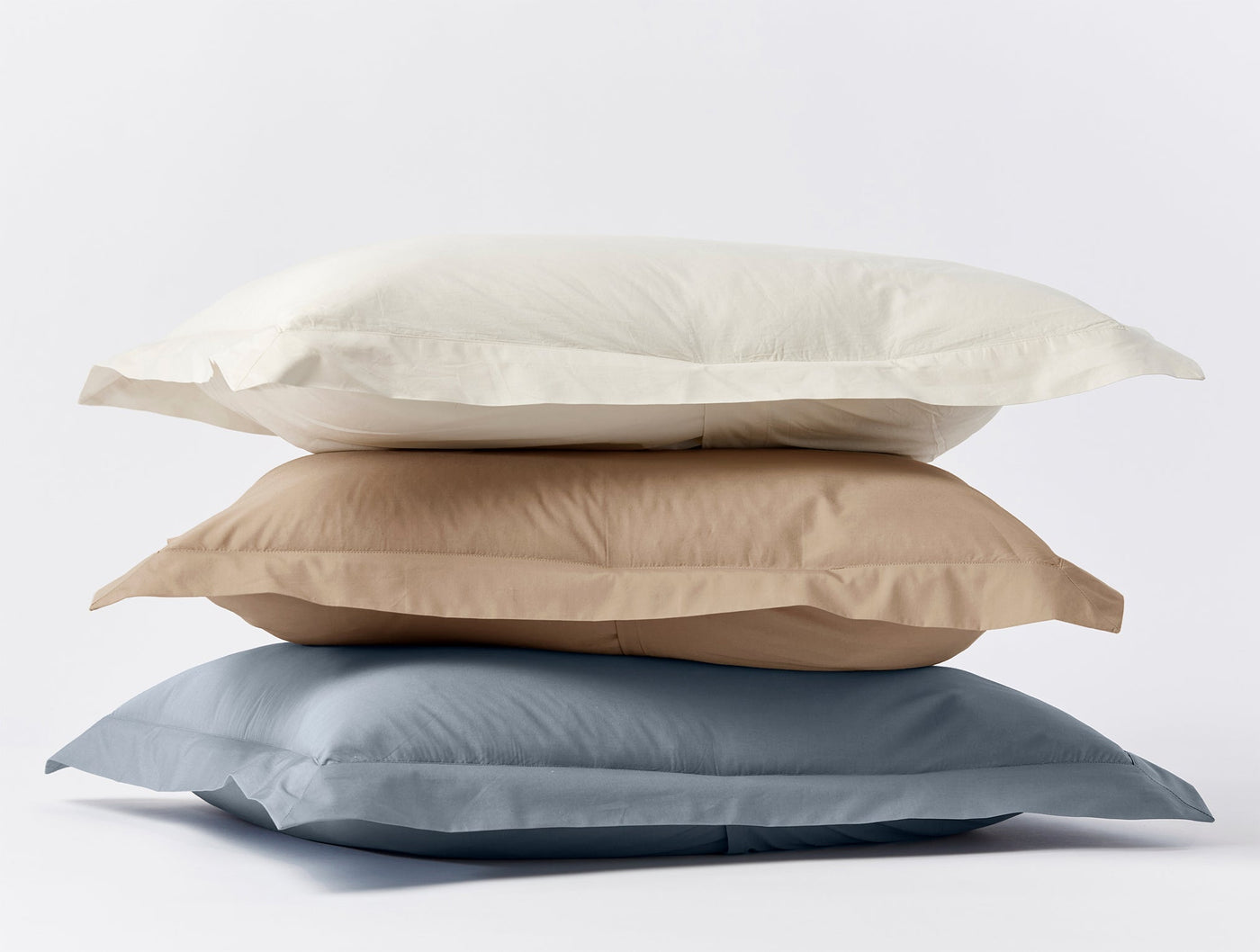 Three stacked Coyuchi Refined Percale shams in ivory, dune, and slate blue.