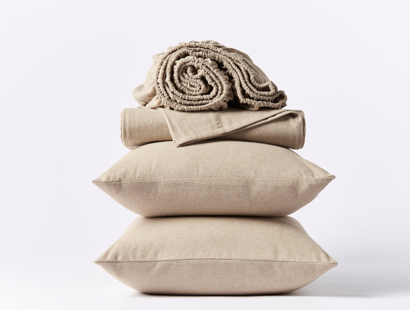 Stack of two sand-colored linen pillows topped with folded flannel sheets and a rolled textured blanket.