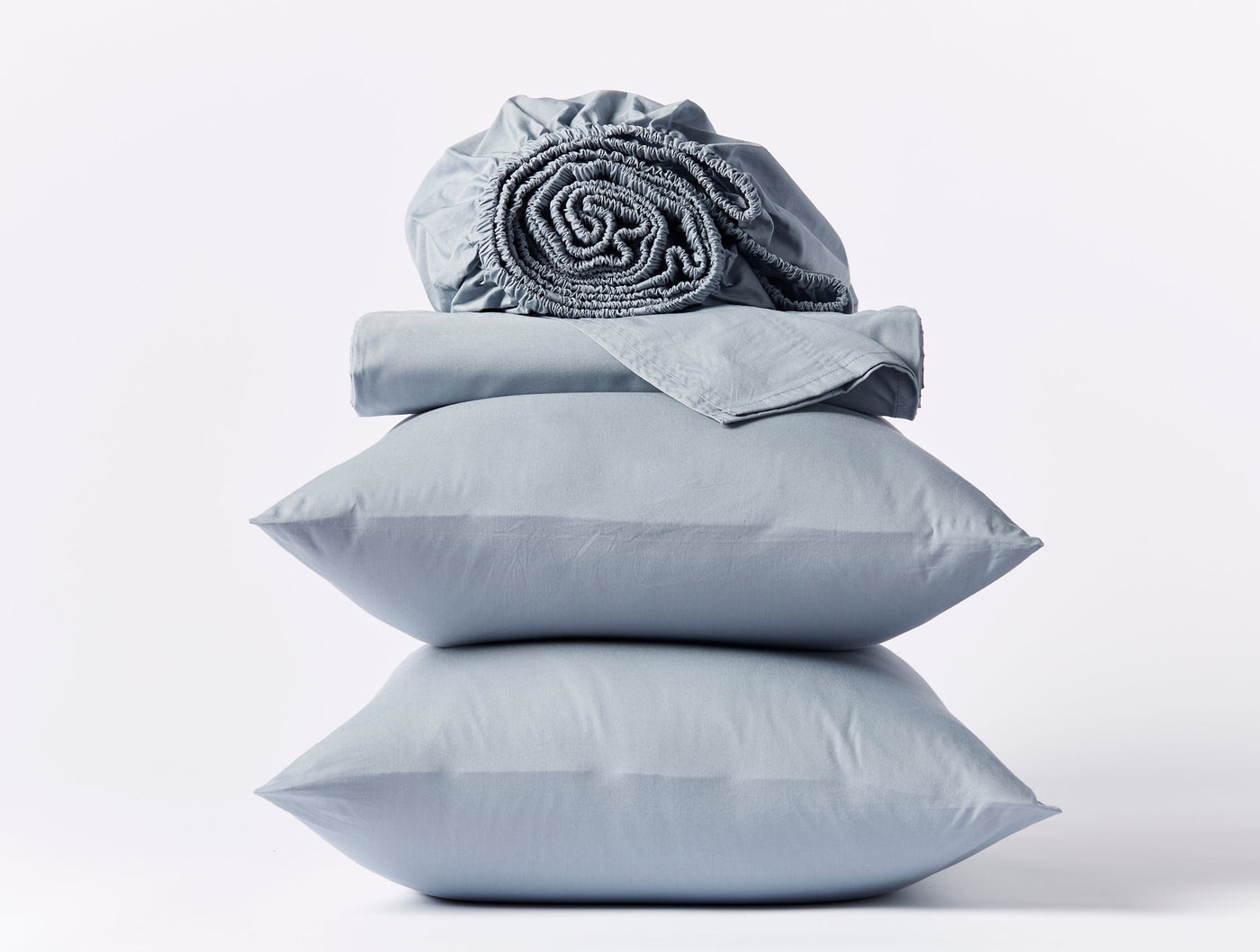 Stacked slate blue Coyuchi percale pillowcases and folded fitted sheet on white background.