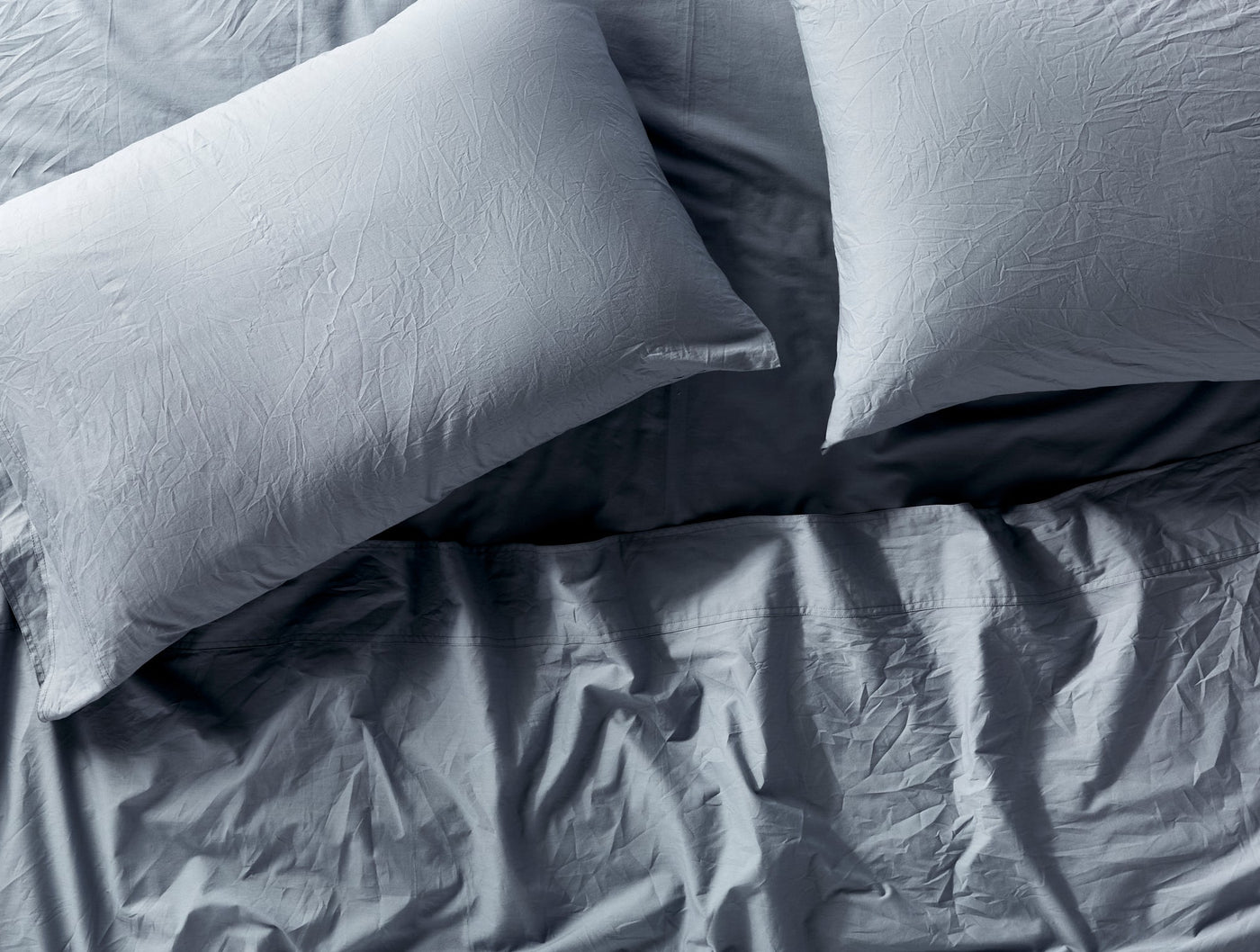 Overhead view of two light blue-grey pillowcases against crinkled percale bedding.