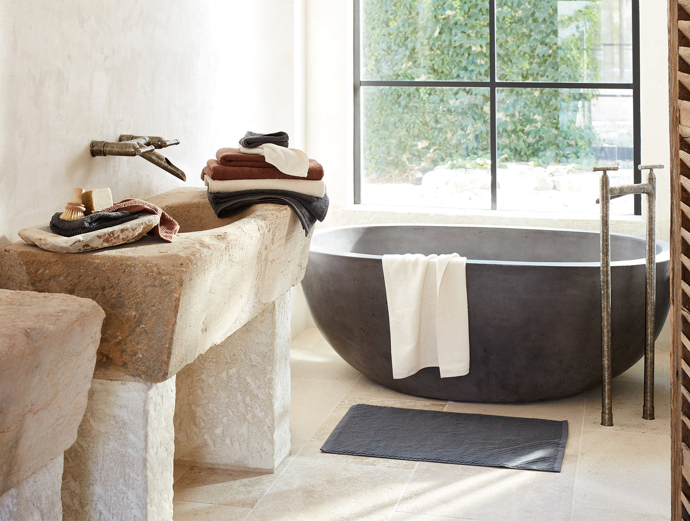 Slate Coyuchi Air Weight bath rug on stone floor beside dark oval soaking tub.