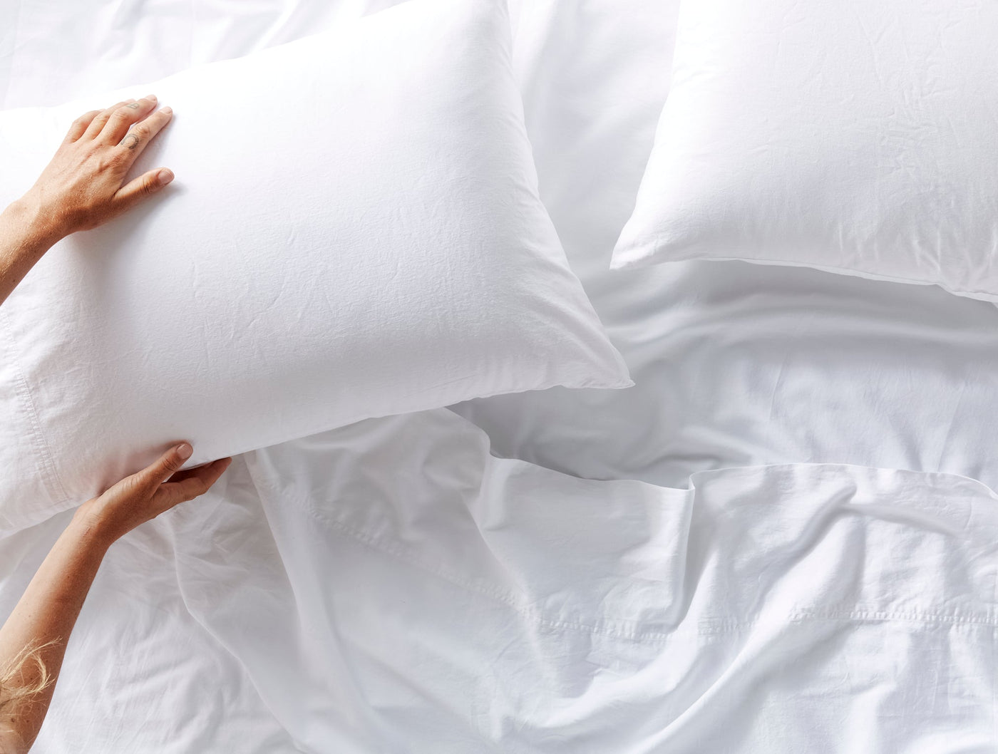 Hands placing a white sateen pillow onto a softly rumpled white bedding arrangement.