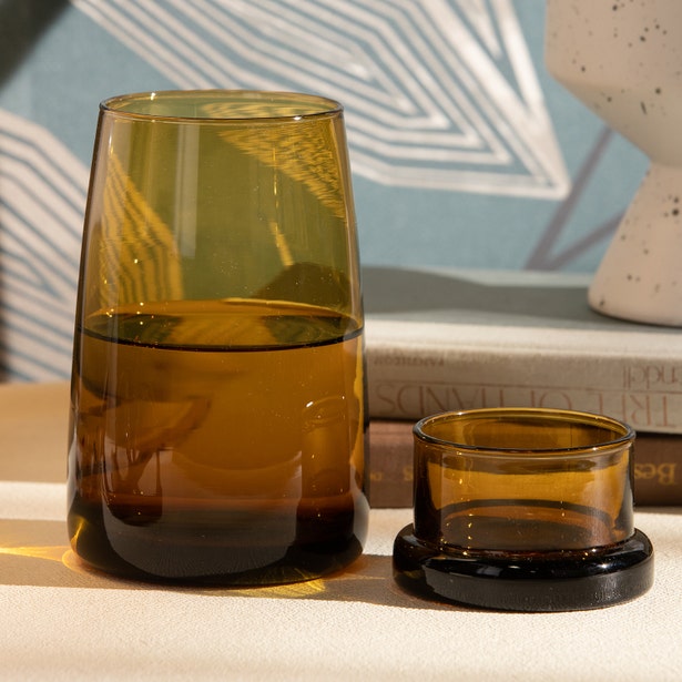 Amber glass carafe with matching cup on dark coaster, styled on a shelf with books.