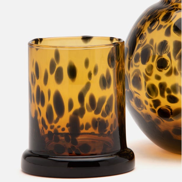 Amber tortoiseshell glass tumbler close-up beside curved carafe, dark spot pattern detail.