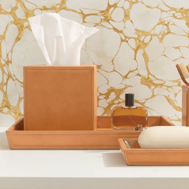 Tan stitched leather bathroom accessories set styled on a white vanity with marble backdrop.