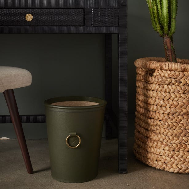 Olive green wastebasket with brass ring handle, styled beside a woven basket near a dark desk.
