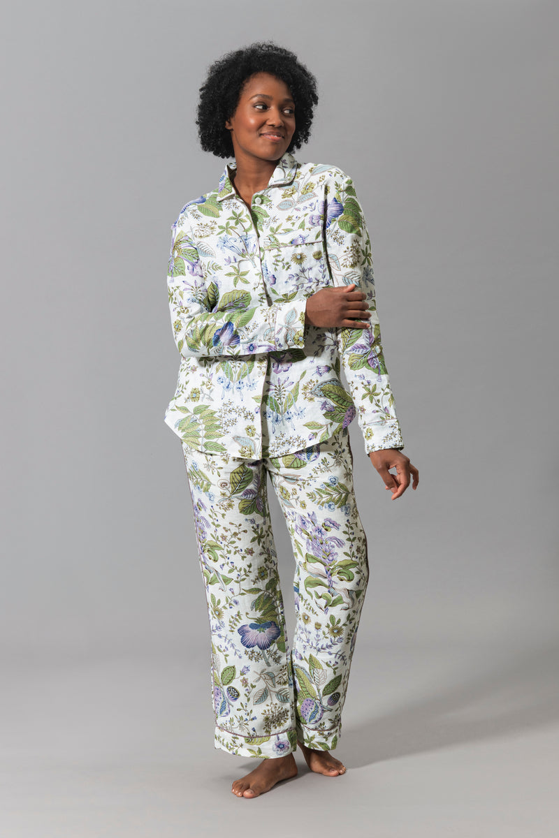A person stands barefoot against a plain gray background, wearing the Matouk Roman Linen Pajama Set in Pomegranate Lilac floral print with matching long-sleeve top and pants.