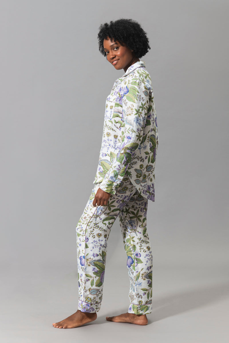 A person stands barefoot on a gray background, wearing the Matouk Roman Linen Pajama Set—a matching long-sleeve, button-up set featuring a Pomegranate Lilac floral pattern.