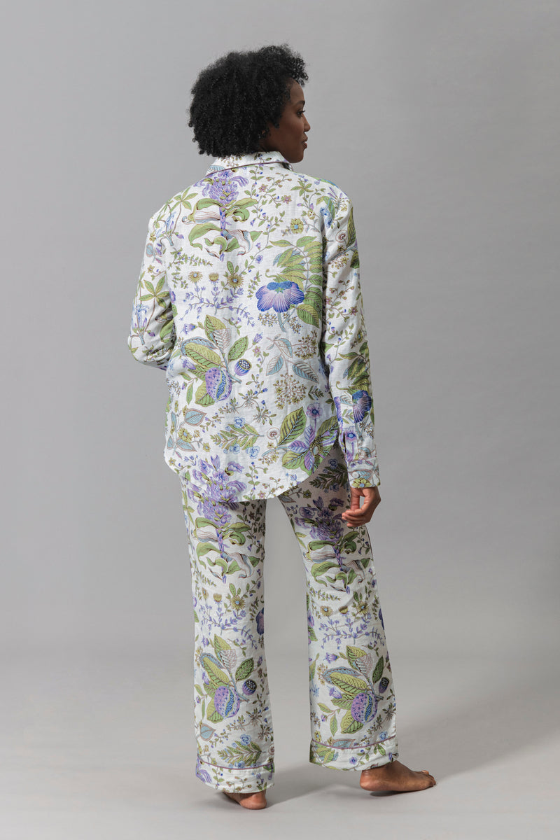 A person stands barefoot facing away, wearing the Matouk Roman Linen Pajama Set featuring green and purple floral patterns against a gray background.