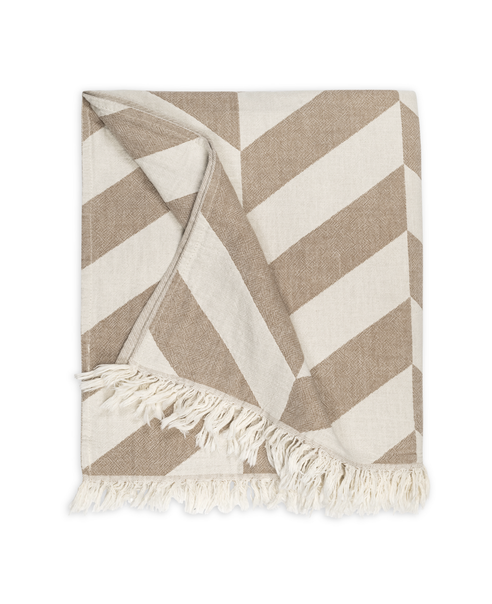 The Matouk Paros Beach Towel features a beige and white diagonal geometric pattern with fringed edges, inspired by classic Turkish bath towels, shown partially folded on a white background.