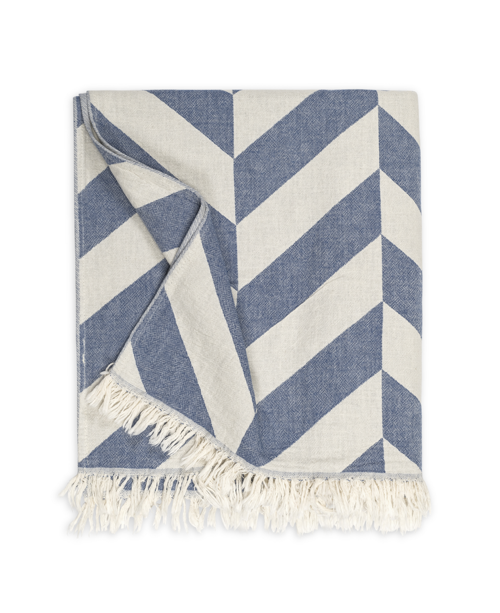 The Paros | Beach Towel by Matouk features a blue and white chevron pattern with fringed edges, shown folded on a plain background.