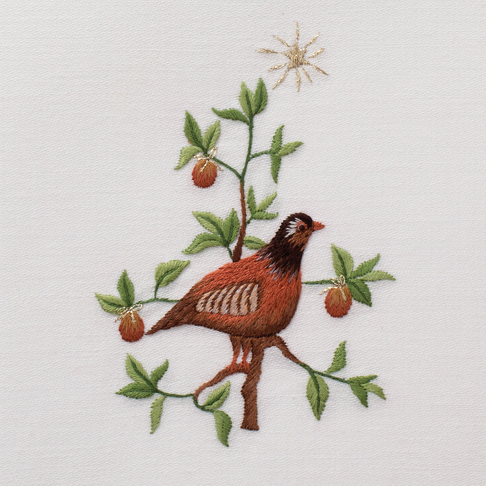 Close-up of embroidered partridge amid green leaves and berries on ivory linen.