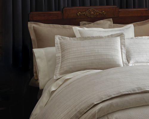A luxurious bed featuring a dark wooden headboard with ornate detailing, dressed in elegant beige, cream, and silver-toned bedding with a subtle striped pattern. Multiple pillows of various sizes and textures, including silk-like and linen finishes, are neatly arranged, enhanced by a book resting on the bed, adding a cozy, inviting touch. The overall atmosphere exudes sophistication and comfort. Featuring Biagio duvet covers by Peacock Alley, available at Linen Society.
