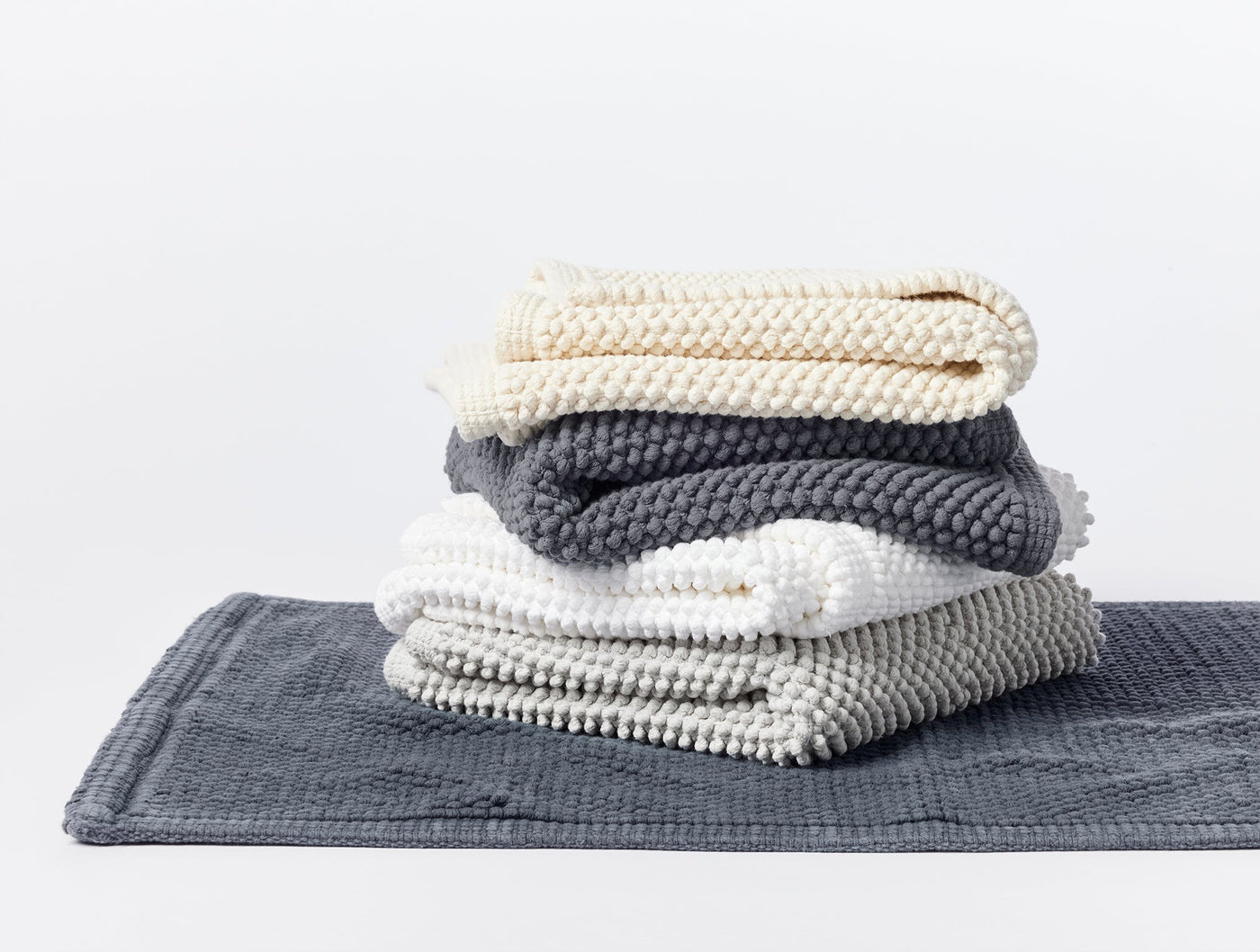 Coyuchi Pebbled bath rugs stacked in ivory, charcoal, white, and slate colorways.