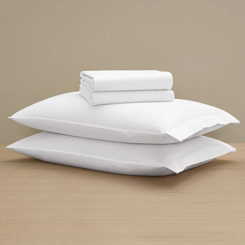 Two Frette Percale Pillow Sham Sets in white are stacked on a wooden surface, topped with a folded 100% cotton duvet cover, all set against a plain beige background.