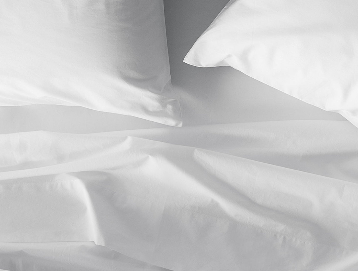 Close-up of crisp white percale sheets with natural folds and two pillows in background.
