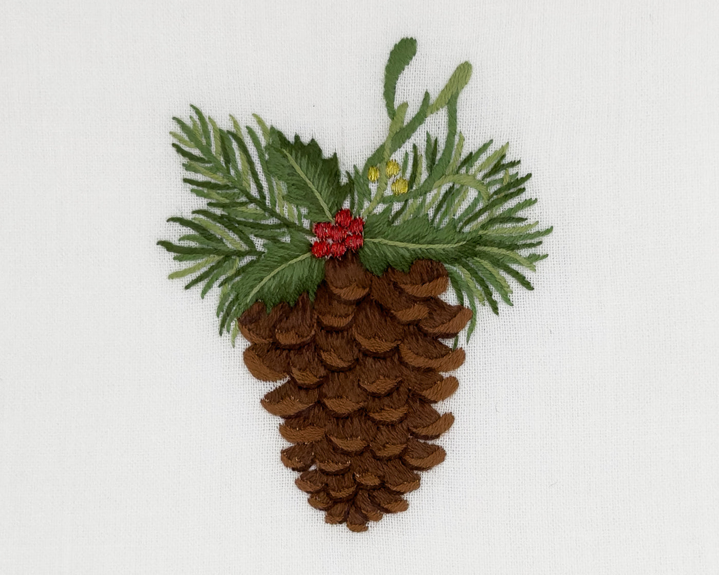 Close-up of embroidered pinecone with holly and red berries on ivory linen.