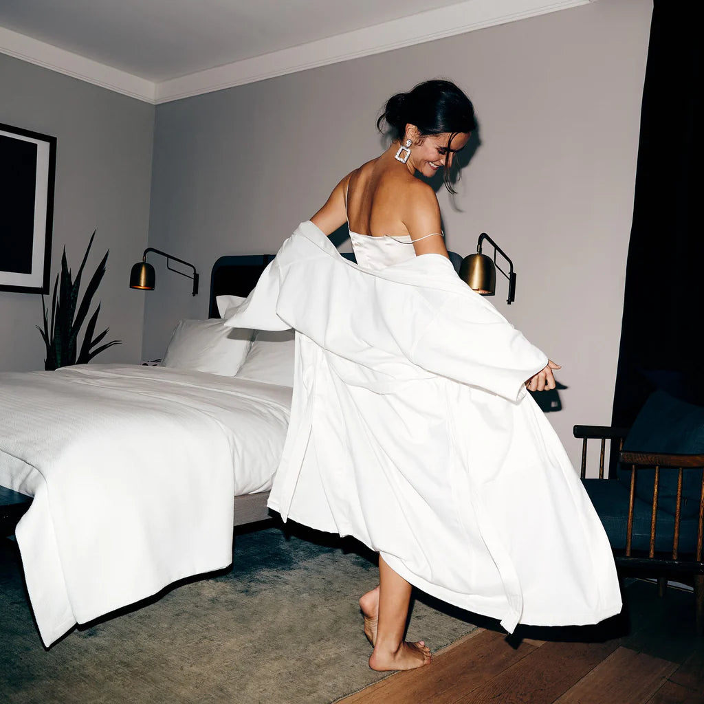 A woman in The Pique Kimono by Frette stands barefoot on a wooden floor near a neatly made bed in a modern bedroom, looking over her shoulder and smiling.