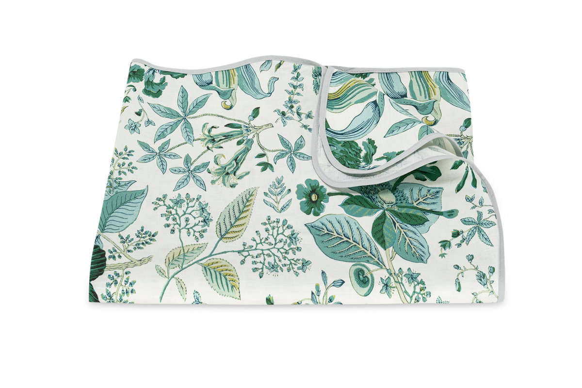 The Matouk Pomegranate Linen Tablecloth features a green and blue botanical leaf and flower print on a white background, finished with a delicate scalloped edge.