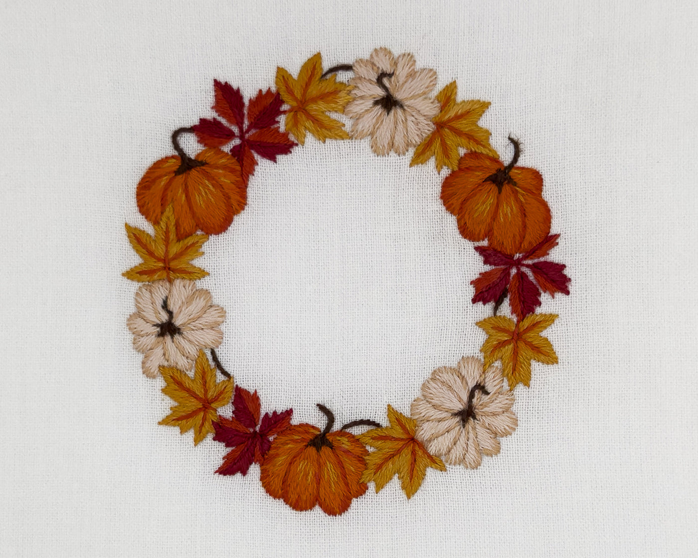 Close-up of embroidered autumn wreath with pumpkins and maple leaves on ivory linen.
