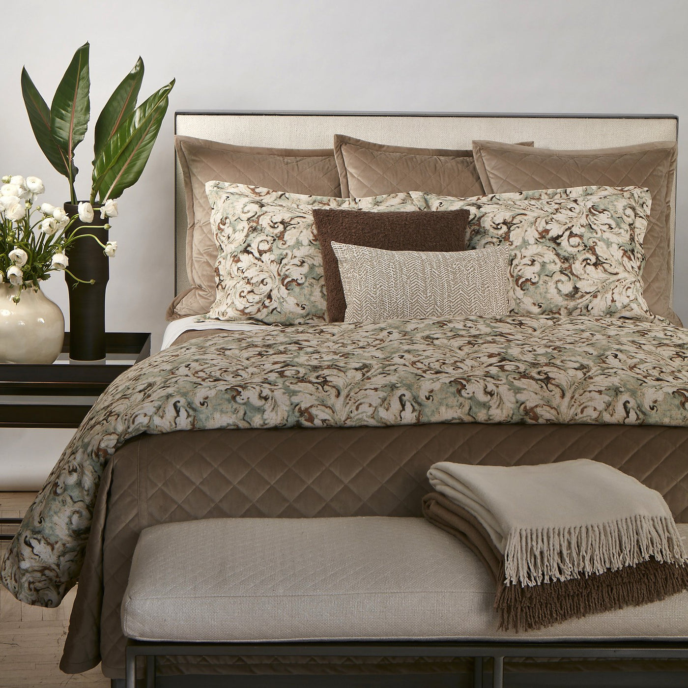 Quilted taupe velvet coverlet styled on bed with floral-print pillows and a folded throw at the foot.